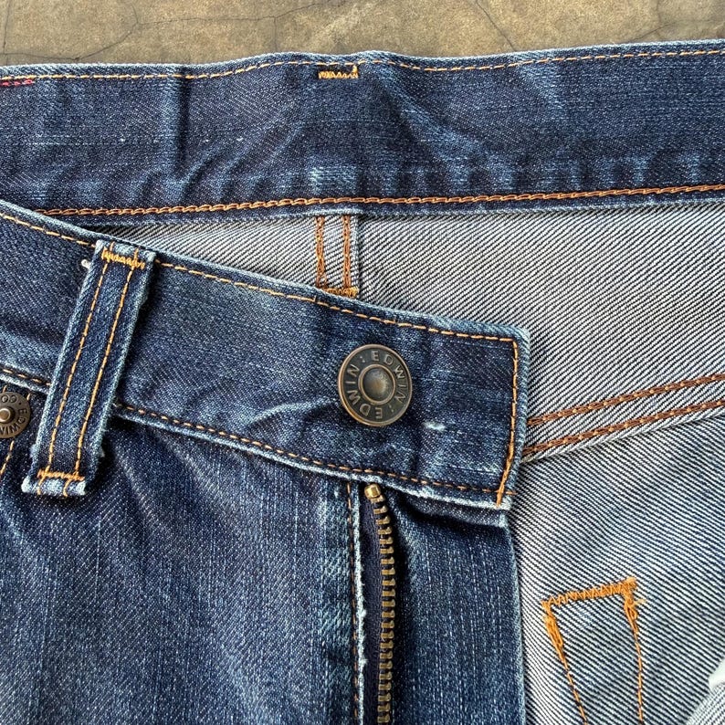 May include: Close-up of a pair of blue denim jeans. The image highlights the details of the jeans, including the zipper, button, and stitching. The button has the brand name "EDWIN" engraved on it.