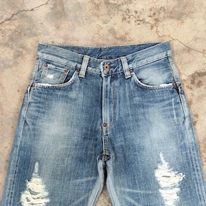 May include: Faded blue denim jeans with distressed details, including ripped sections on the legs. The jeans feature classic five-pocket styling, a button closure, and belt loops. The denim has a worn, vintage look.
