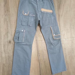 Puede incluir: Pantalones cargo grises con múltiples bolsillos y cremalleras. Los pantalones tienen un diseño de pierna recta y detalles en color beige en los bolsillos. Los pantalones están colgados en una percha sobre un fondo de madera.