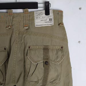 May include: Khaki-colored utility trousers with multiple pockets and a button closure. The back of the pants features a label with the text "XU" and other details. The stitching is in a contrasting orange color.