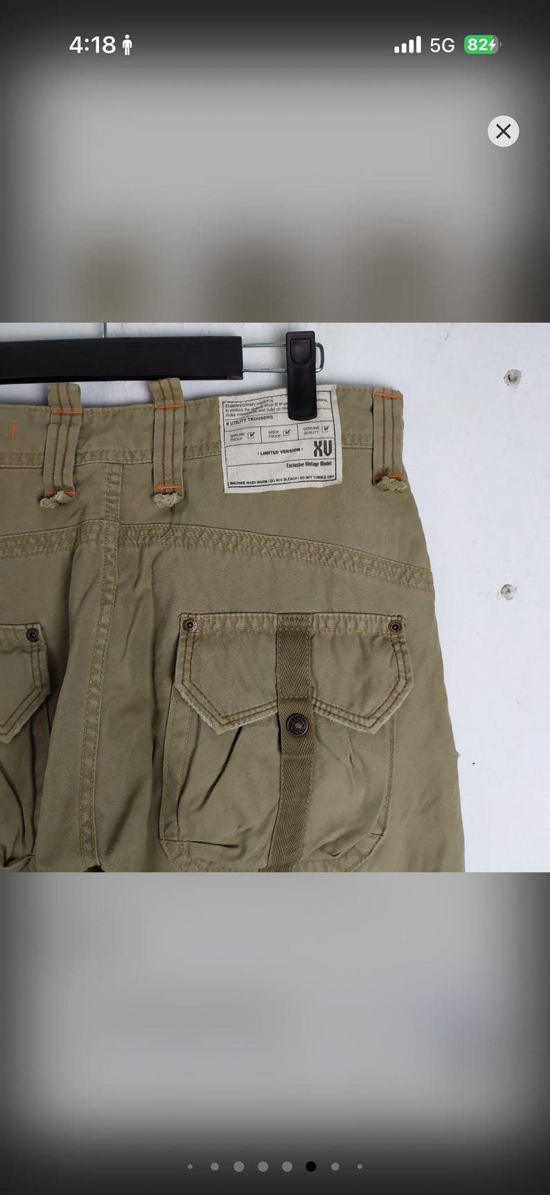 May include: Khaki-colored utility trousers with multiple pockets and a button closure. The back of the pants features a label with the text "XU" and other details. The stitching is in a contrasting orange color.