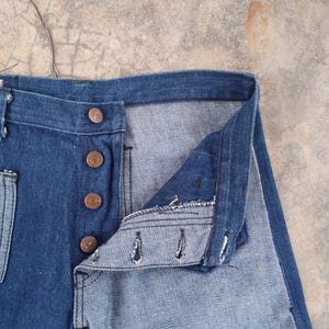 May include: Close-up of blue denim jeans with a button fly. The jeans have copper-colored buttons and a classic five-pocket design. The denim fabric shows signs of wear, suggesting a vintage or well-loved item. The jeans are unbuttoned, revealing the interior.