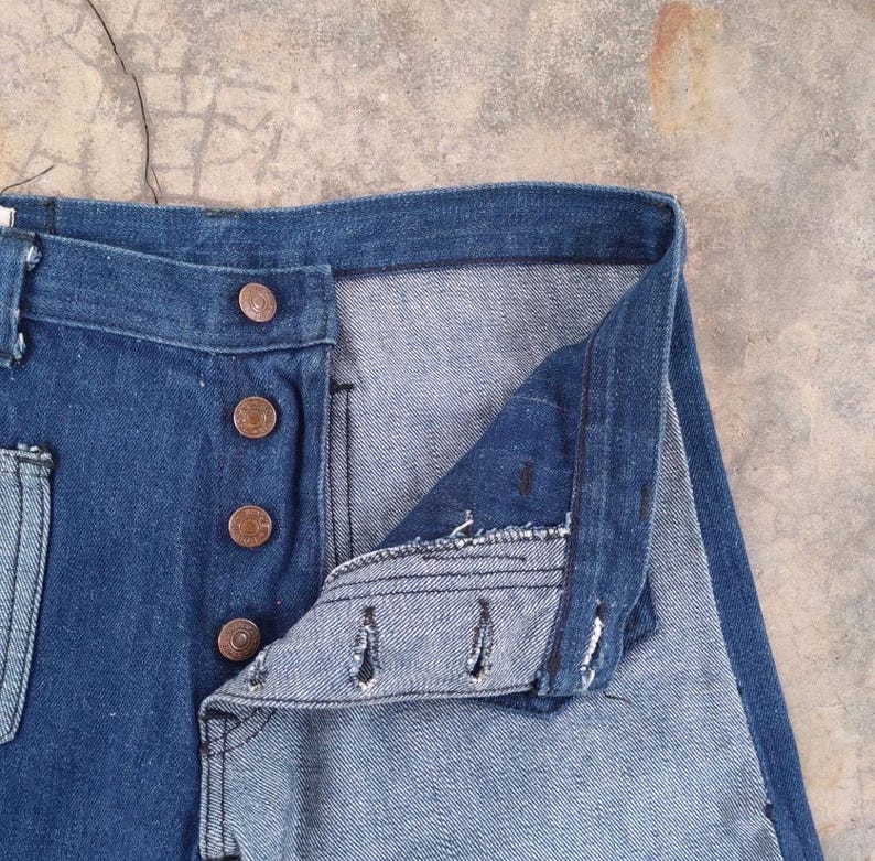 May include: Close-up of blue denim jeans with a button fly. The jeans have copper-colored buttons and a classic five-pocket design. The denim fabric shows signs of wear, suggesting a vintage or well-loved item. The jeans are unbuttoned, revealing the interior.