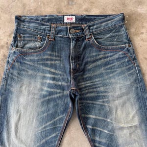 Pu&ograve; includere: Primo piano di jeans di denim blu con un lavaggio sbiadito e dettagli usurati. I jeans presentano un classico design a cinque tasche, una chiusura con bottone e cerniera e cuciture rosse. Il marchio "EDWIN" &egrave; visibile su una piccola etichetta.