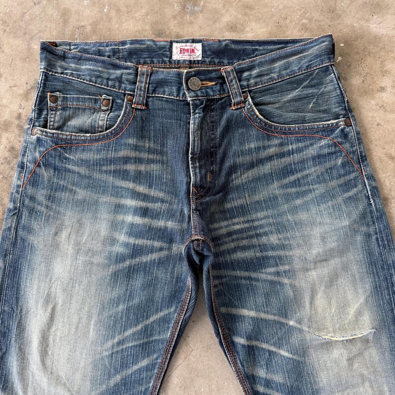 Pu&ograve; includere: Primo piano di jeans di denim blu con un lavaggio sbiadito e dettagli usurati. I jeans presentano un classico design a cinque tasche, una chiusura con bottone e cerniera e cuciture rosse. Il marchio "EDWIN" &egrave; visibile su una piccola etichetta.