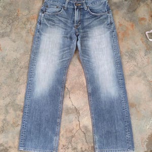 May include: Light blue denim jeans with a faded wash effect. The jeans have a classic five-pocket design, a button and zipper closure, and a straight leg fit. The denim shows wear and tear, with fading and subtle distressing.