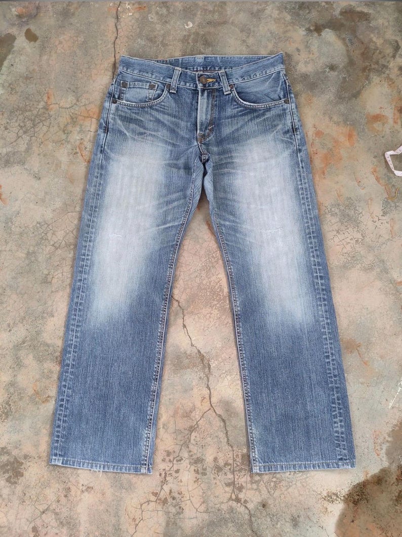 May include: Light blue denim jeans with a faded wash effect. The jeans have a classic five-pocket design, a button and zipper closure, and a straight leg fit. The denim shows wear and tear, with fading and subtle distressing.