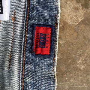 May include: Close-up of denim jeans with a red rectangular patch. The patch features the number "503" and the brand name "Edwin" in white text. The jeans have visible stitching and a textured appearance.
