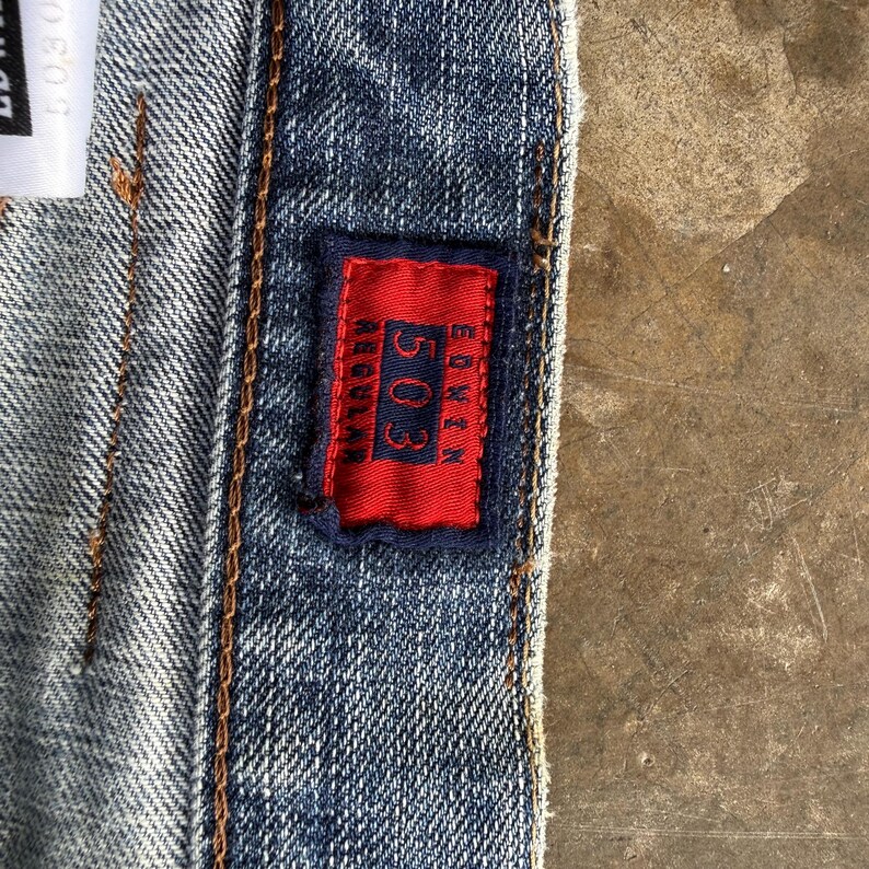 May include: Close-up of denim jeans with a red rectangular patch. The patch features the number "503" and the brand name "Edwin" in white text. The jeans have visible stitching and a textured appearance.