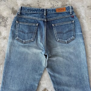 May include: Faded blue denim jeans with a high waist and two back pockets. The jeans have a brown leather patch with the word "SOMETHING" on the back waistband. The denim has a worn, vintage look.