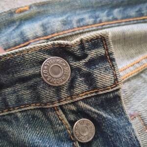 May include: Close-up of a section of blue denim jeans. The jeans have a worn, faded appearance with visible stitching in orange thread. Two silver metal buttons are visible, each with the text "JOE McCOY & CO." around the edge.