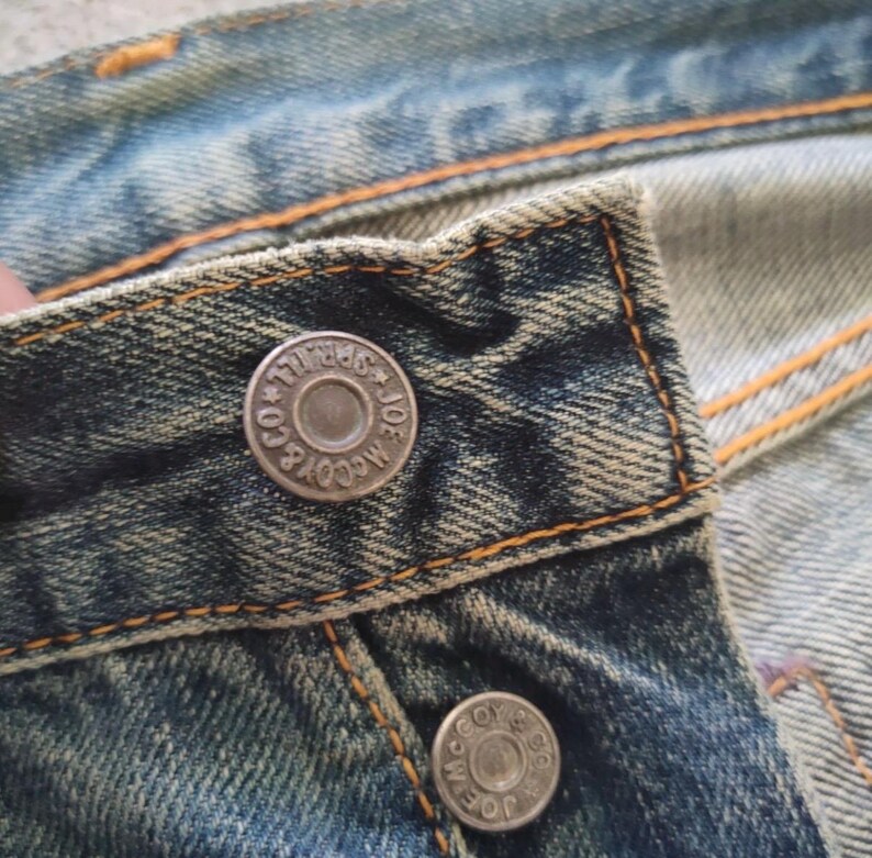 May include: Close-up of a section of blue denim jeans. The jeans have a worn, faded appearance with visible stitching in orange thread. Two silver metal buttons are visible, each with the text "JOE McCOY & CO." around the edge.