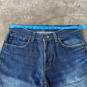 May include: Close-up of a pair of dark blue denim jeans. The jeans have a classic five-pocket design, a button closure, and a zipper fly. A blue measuring tape is laid across the top of the jeans, measuring approximately 17 inches across.