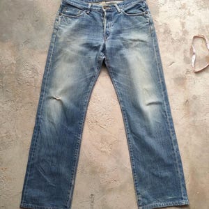 May include: Light blue denim jeans with a classic five-pocket design. The jeans show signs of wear with faded areas and a small tear on the left knee. A measuring tape is visible in the background.