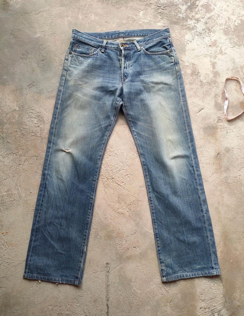 May include: Light blue denim jeans with a classic five-pocket design. The jeans show signs of wear with faded areas and a small tear on the left knee. A measuring tape is visible in the background.