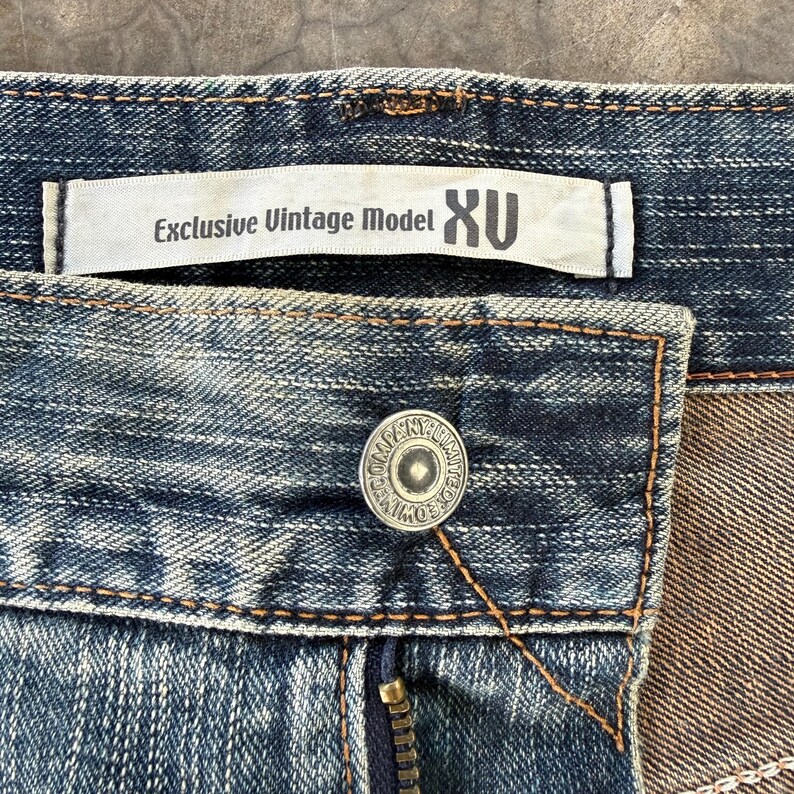Puede incluir: Primer plano de unos vaqueros azules con un bot&oacute;n y cremallera. Los vaqueros tienen una etiqueta que dice "Exclusive Vintage Model XU". El bot&oacute;n es plateado con el texto "COMPANY LIMITED".