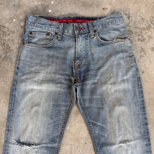 May include: Light blue denim jeans with a faded wash and distressed details. Features classic five-pocket styling, a button and zipper closure, and red stitching accents. The jeans have a ripped knee and a red waistband label that reads "EDGE on Blue Jeans".