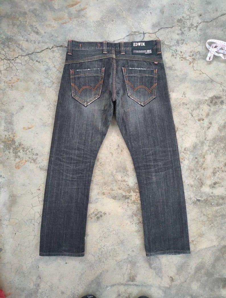 Pu&ograve; includere: Jeans in denim grigio scuro con cuciture arancioni. I jeans presentano due tasche posteriori con un design unico e il marchio "EDWIN" sul retro. I jeans sono appoggiati su una superficie di cemento.