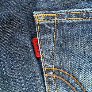 May include: Close-up of a section of blue denim jeans. The back pocket is visible, with orange stitching and a small red tag that reads "EDWIN". The denim has a textured appearance.