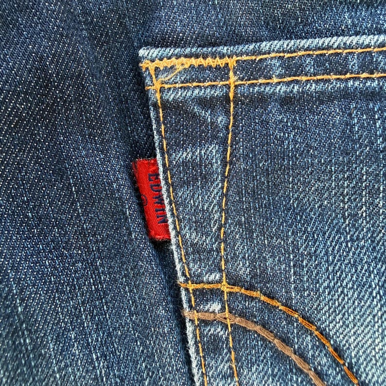 May include: Close-up of a section of blue denim jeans. The back pocket is visible, with orange stitching and a small red tag that reads "EDWIN". The denim has a textured appearance.