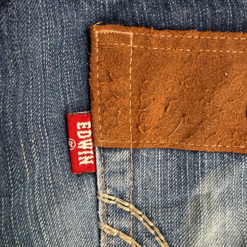 Op de afbeelding: Close-up van een blauwe spijkerbroek met een bruine leren patch en een rood label. Het rode label heeft de merknaam "Edwin" in witte letters. De leren patch is op de jeans gestikt.