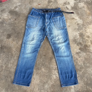 May include: Blue denim pants with large front pockets and a black belt. The pants have a relaxed fit and a faded wash, with some distressed details. The waistband is elasticized. The pants are laid flat on a concrete surface.