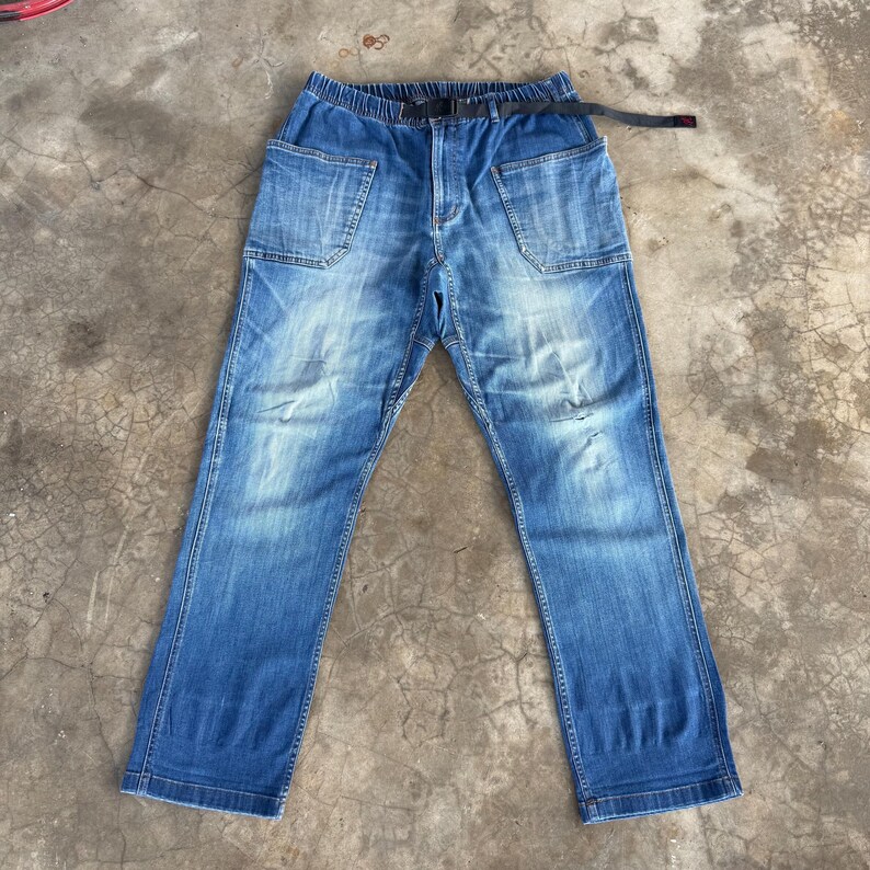 May include: Blue denim pants with large front pockets and a black belt. The pants have a relaxed fit and a faded wash, with some distressed details. The waistband is elasticized. The pants are laid flat on a concrete surface.