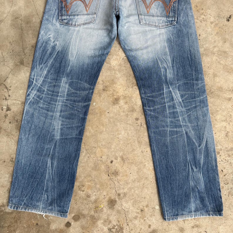 May include: Faded blue denim jeans with a distressed appearance. The jeans feature a classic five-pocket design, with a light wash on the upper portion and darker shades towards the bottom. The back pockets have a distinctive orange stitching.