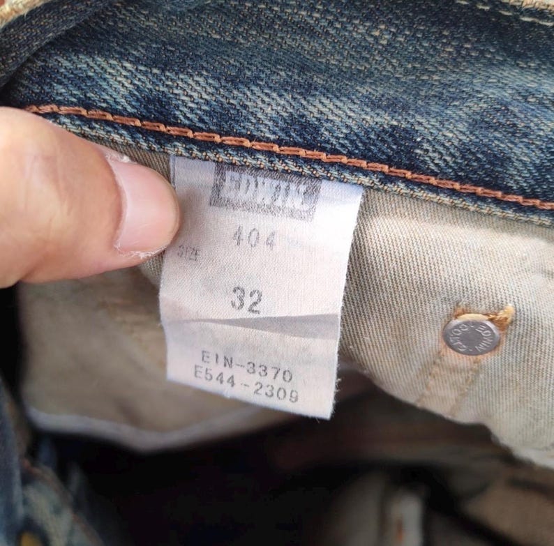May include: Close-up of a denim jeans label with the brand name "EDWIN" at the top. The label shows the size "32" and style number "404". Additional numbers and letters are also visible on the label.
