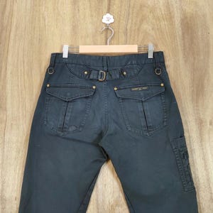 Pu&ograve; includere: Pantaloni cargo blu navy scuro con tasche multiple e passanti per cintura. I pantaloni hanno una chiusura con fibbia e rivetti color oro. Il marchio "EDWIN GOLF" &egrave; visibile sulla tasca posteriore. I pantaloni sono appesi a una gruccia di legno.