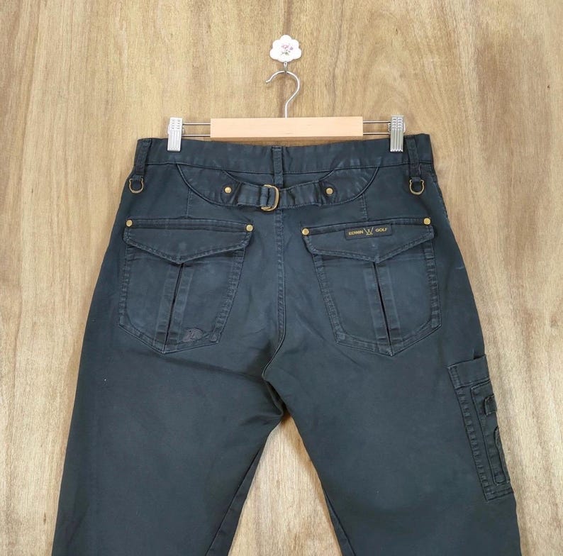 Pu&ograve; includere: Pantaloni cargo blu navy scuro con tasche multiple e passanti per cintura. I pantaloni hanno una chiusura con fibbia e rivetti color oro. Il marchio "EDWIN GOLF" &egrave; visibile sulla tasca posteriore. I pantaloni sono appesi a una gruccia di legno.
