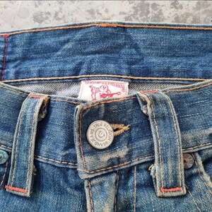 May include: Close-up of blue denim jeans with orange stitching. The jeans feature a button closure with the text "TRUE RELIGION" and a brand label. The denim has a worn, faded appearance.