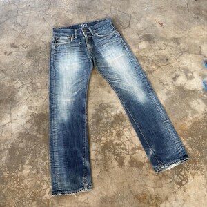 May include: Blue denim jeans with a faded wash and distressed details. The jeans have a classic five-pocket design, a button fly, and the number "503" is visible on the waistband. The jeans are laid flat on a concrete surface.