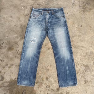 May include: Light blue denim jeans with a faded wash and distressed details. The jeans have a classic five-pocket design, a button and zipper closure, and a straight leg fit. The denim has a worn appearance with subtle fading and a few small tears.