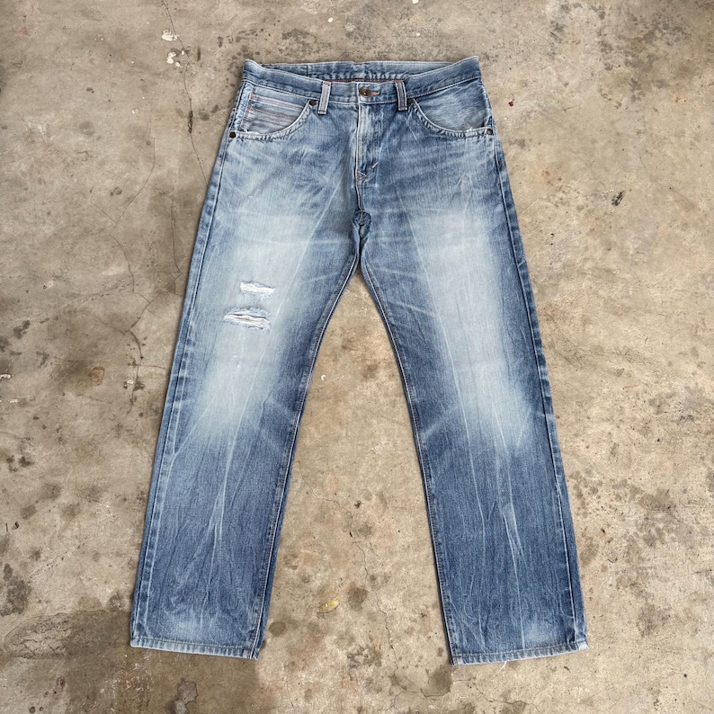 May include: Light blue denim jeans with a faded wash and distressed details. The jeans have a classic five-pocket design, a button and zipper closure, and a straight leg fit. The denim has a worn appearance with subtle fading and a few small tears.