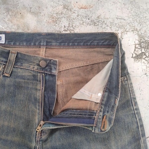 May include: Close-up of blue denim jeans with a partially unzipped fly. The jeans have a button closure and a classic five-pocket design. The interior lining is a contrasting tan color, and the denim shows signs of wear.