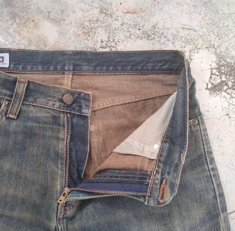 May include: Close-up of blue denim jeans with a partially unzipped fly. The jeans have a button closure and a classic five-pocket design. The interior lining is a contrasting tan color, and the denim shows signs of wear.