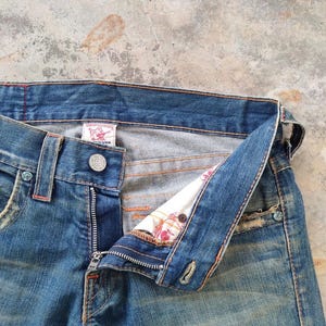 May include: Close-up of blue denim jeans with an open fly. The jeans feature a button closure, belt loops, and a small label. The interior lining, with a floral pattern, is visible. The denim has a faded, worn look, with visible stitching and detailing.