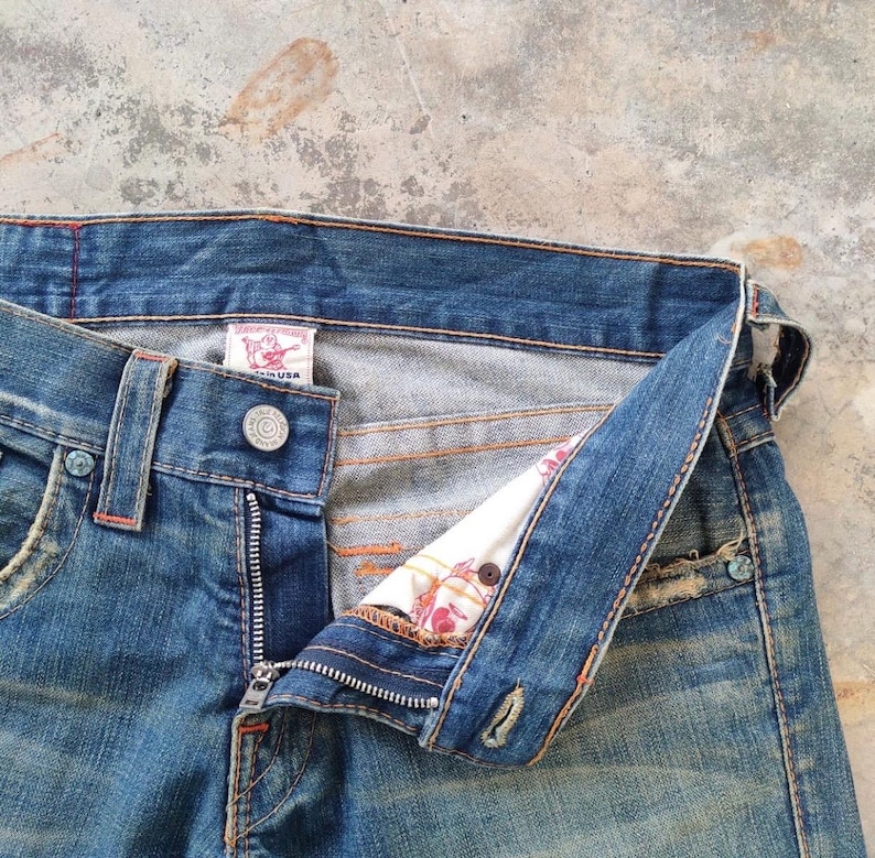 May include: Close-up of blue denim jeans with an open fly. The jeans feature a button closure, belt loops, and a small label. The interior lining, with a floral pattern, is visible. The denim has a faded, worn look, with visible stitching and detailing.