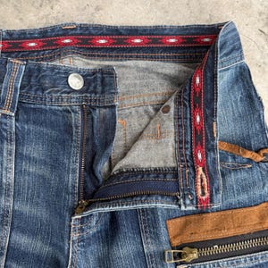 Op de afbeelding: Close-up van een blauwe spijkerbroek met een gedeeltelijk open rits. De tailleband heeft een decoratief zwart-rood patroonlint. Een bruin lederen accent en een rits detailleren de voorkant van de jeans. De jeans heeft een knoopsluiting en een zilveren knoop.