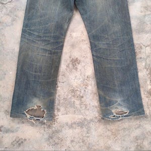 May include: Faded blue denim jeans with distressed details. The jeans show wear and tear, with visible fading and holes near the ankles. The fabric has a worn appearance, suggesting a vintage or well-loved item.