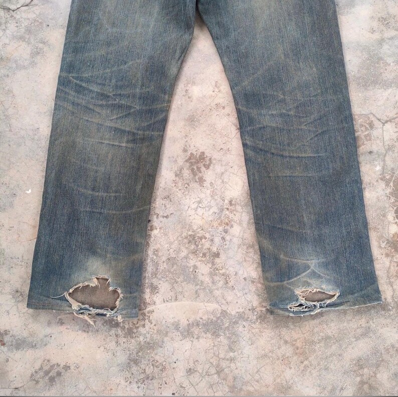 May include: Faded blue denim jeans with distressed details. The jeans show wear and tear, with visible fading and holes near the ankles. The fabric has a worn appearance, suggesting a vintage or well-loved item.