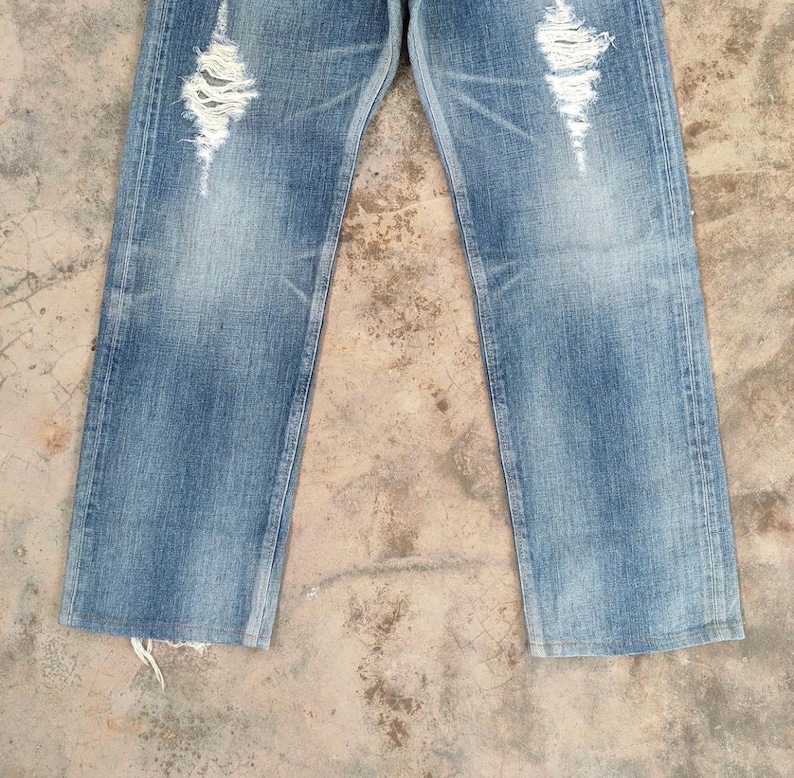 May include: Light blue denim jeans with distressed detailing. The jeans feature ripped sections on the front of the legs, creating a worn and vintage aesthetic. The denim has a faded appearance, adding to the casual style.