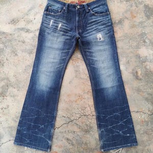 May include: Blue denim jeans with a faded wash and distressed details. The jeans have a classic five-pocket design, a button and zipper closure, and a flared leg. The waistband features a patterned lining.