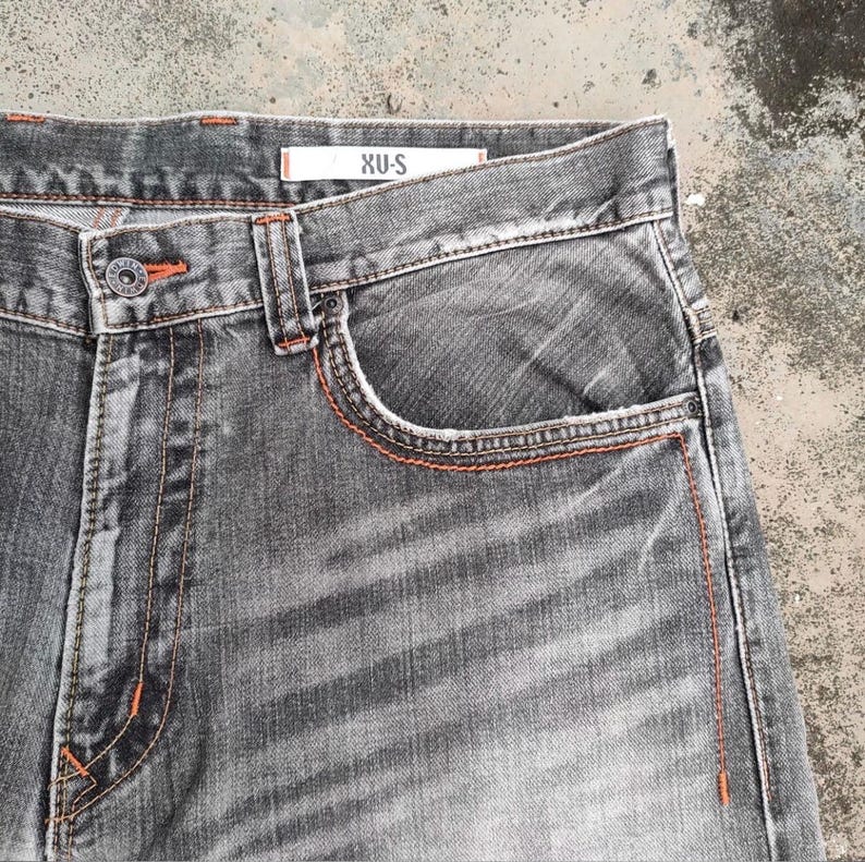 K&ouml;nnte beinhalten: Nahaufnahme einer grauen Jeans aus Denim mit orangefarbenen N&auml;hten. Die Jeans hat ein klassisches F&uuml;nf-Taschen-Design, einen Knopfverschluss und den Markennamen "XU-S" auf dem Etikett. Der Stoff hat einen verblichenen, abgenutzten Look.
