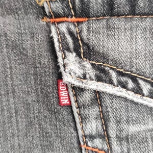 K&ouml;nnte beinhalten: Nahaufnahme von grauem Denim-Stoff mit orangefarbenen N&auml;hten und einem roten Edwin-Etikett. Der Denim hat ein abgenutztes Aussehen mit sichtbaren N&auml;hten und einer strukturierten Oberfl&auml;che. Die N&auml;hte setzen einen farblichen Kontrast zum grauen Denim.