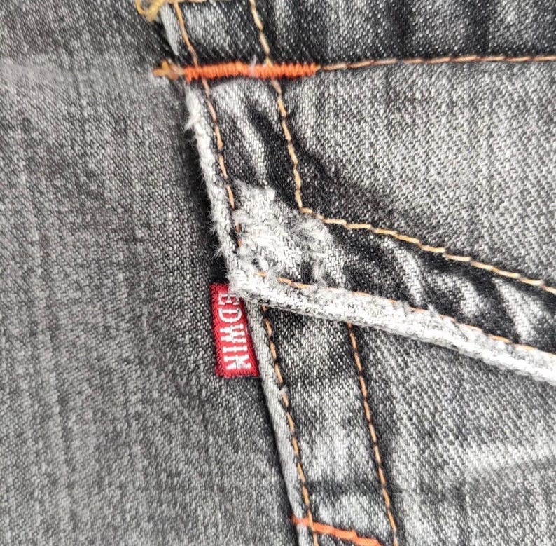 K&ouml;nnte beinhalten: Nahaufnahme von grauem Denim-Stoff mit orangefarbenen N&auml;hten und einem roten Edwin-Etikett. Der Denim hat ein abgenutztes Aussehen mit sichtbaren N&auml;hten und einer strukturierten Oberfl&auml;che. Die N&auml;hte setzen einen farblichen Kontrast zum grauen Denim.