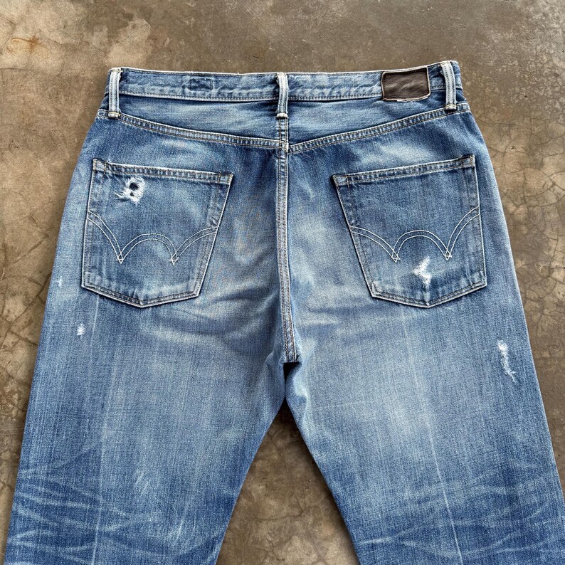 Pu&ograve; includere: Jeans in denim blu chiaro con un aspetto usurato, che mostra segni di usura. Le tasche posteriori hanno un caratteristico design con cuciture curve. Una toppa in pelle marrone &egrave; visibile in vita. I jeans hanno un classico design a cinque tasche.