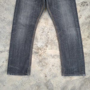 Pu&ograve; includere: Un paio di jeans di denim grigio scuro. I jeans hanno un taglio dritto e presentano cuciture visibili in un colore beige a contrasto lungo le cuciture. Il tessuto appare usurato, con una sottile sbiaditura.