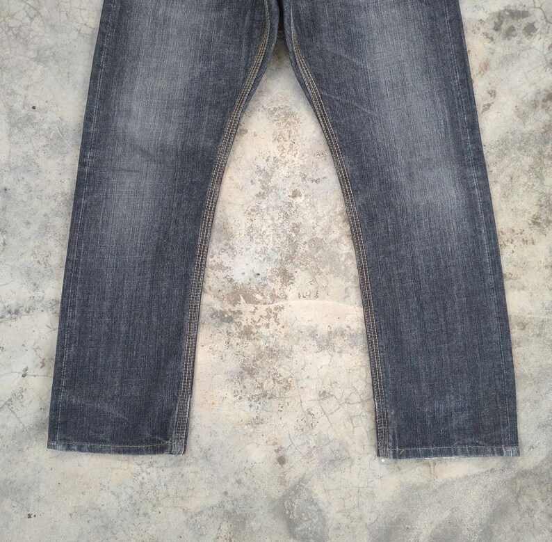 Pu&ograve; includere: Un paio di jeans di denim grigio scuro. I jeans hanno un taglio dritto e presentano cuciture visibili in un colore beige a contrasto lungo le cuciture. Il tessuto appare usurato, con una sottile sbiaditura.